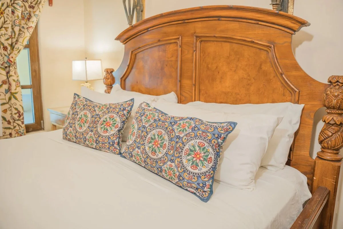 A neatly made bed with white bedding, decorative pillows with floral patterns, a wooden headboard, a nightstand, a lamp, and floral curtains in a cozy bedroom.