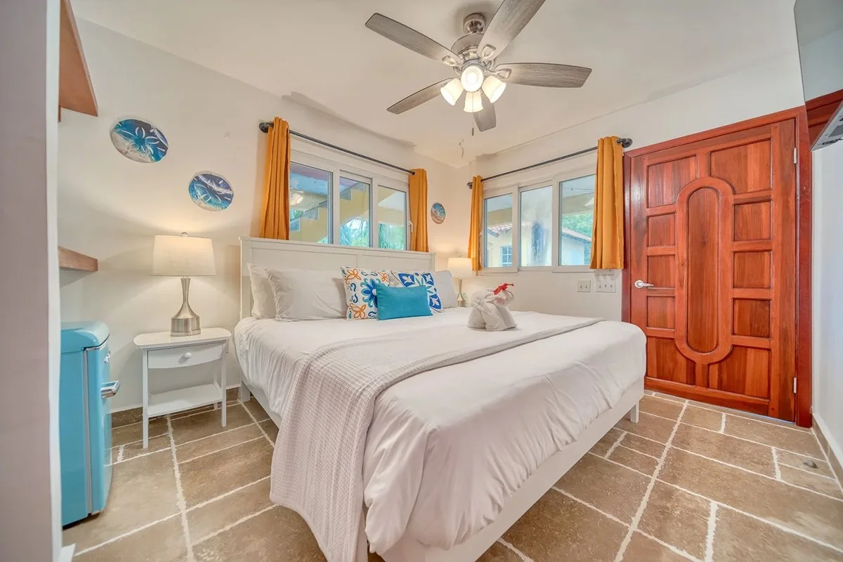 A bedroom with a large bed, white bedding, colorful pillows, two nightstands with lamps, and windows with yellow curtains. There's a wooden door and a ceiling fan with lights.
