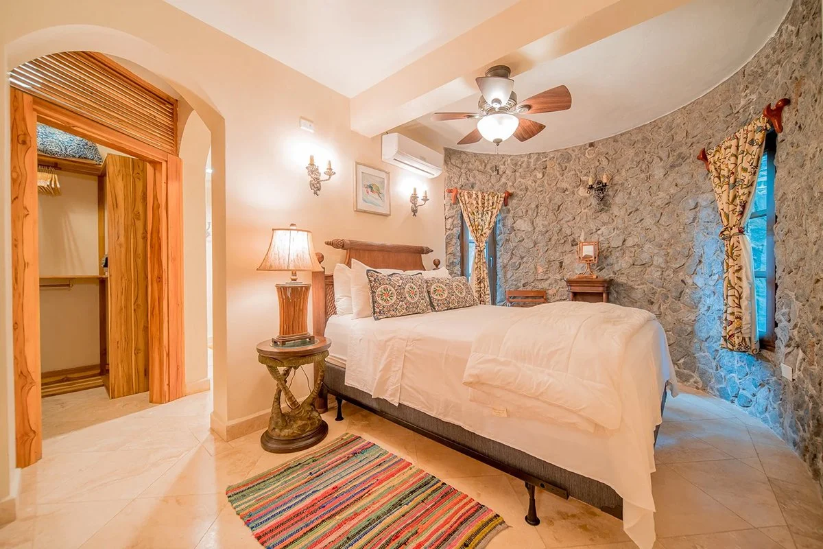 Bedroom with stone accent wall, white bed with decorative pillows, wooden furniture, colorful window curtains, ceiling fan, and a vibrant striped rug.