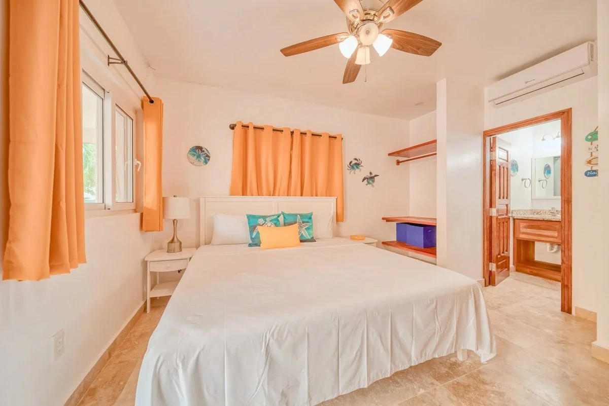 A bedroom with a white bed, orange curtains, a ceiling fan with wooden blades, and wall decorations of sea creatures, with a small bathroom visible through an open door.