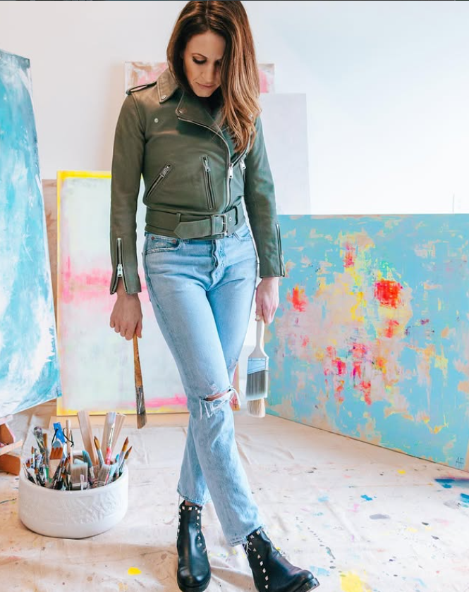Woman in olive green jacket and ripped jeans standing in an art studio, holding paintbrushes, surrounded by abstract paintings and art supplies.