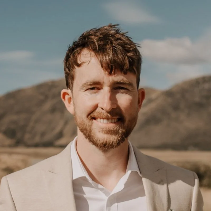 A smiling man with brown hair and a beard, wearing a light-colored suit jacket and white shirt, standing outdoors with hills and a partly cloudy sky in the background.