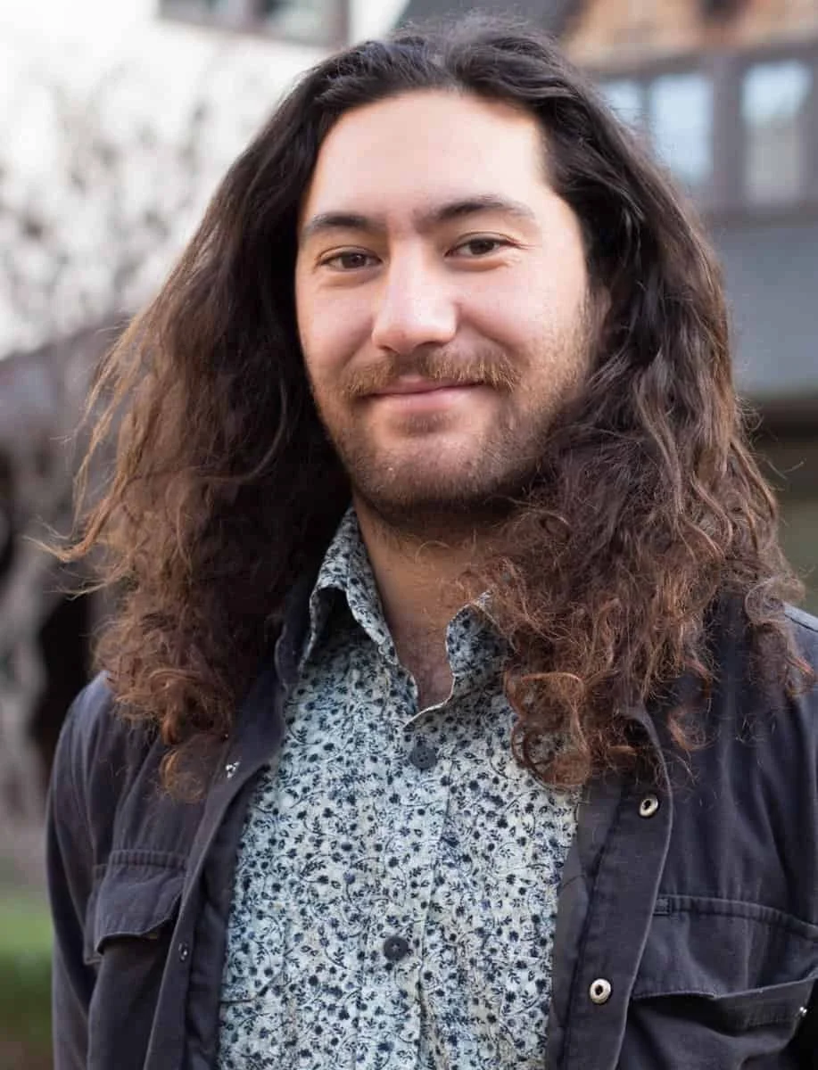 A man with long, curly brown hair and a beard, wearing a patterned shirt and a dark jacket, smiling outdoors.