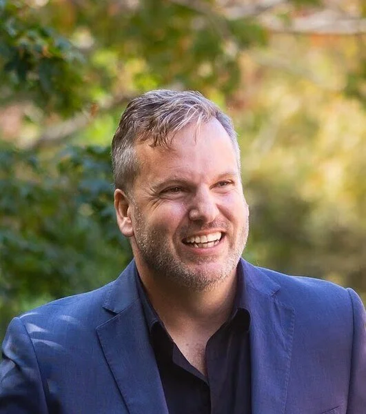 A man with light brown hair and a beard, smiling outdoors, wearing a blue suit jacket and black shirt with a blurred green and autumn-colored background.