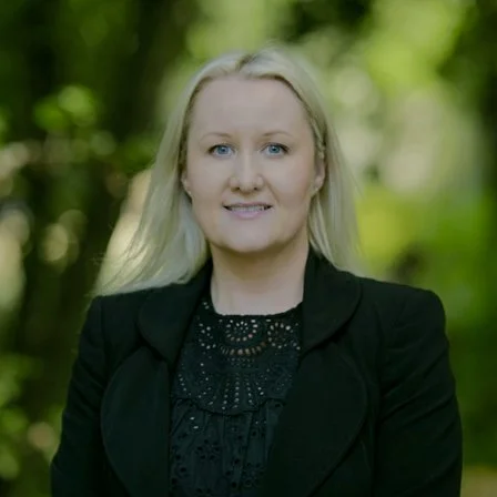 A woman with long blonde hair smiling outdoors in a green park, wearing a black blazer and a black top.