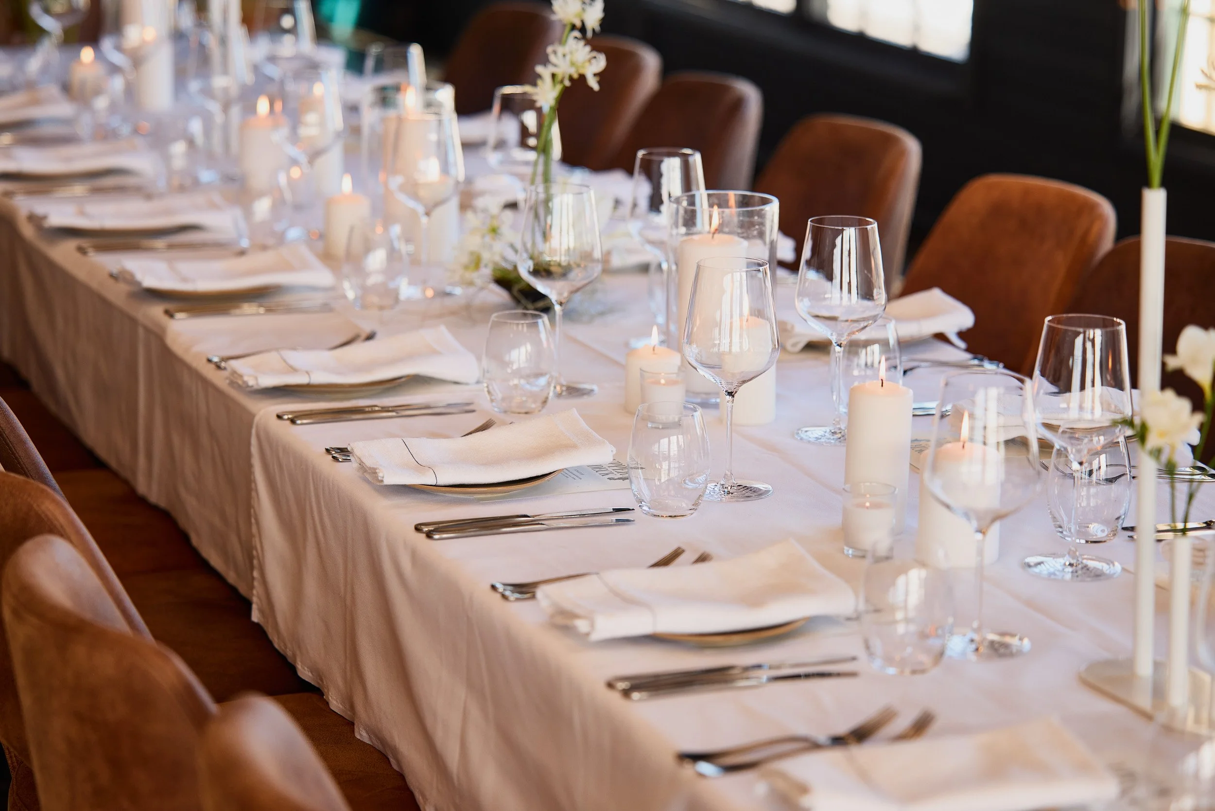 Elegant dining table set with white tablecloth, wine glasses, candles, and floral centerpiece in a warm-lit room.
