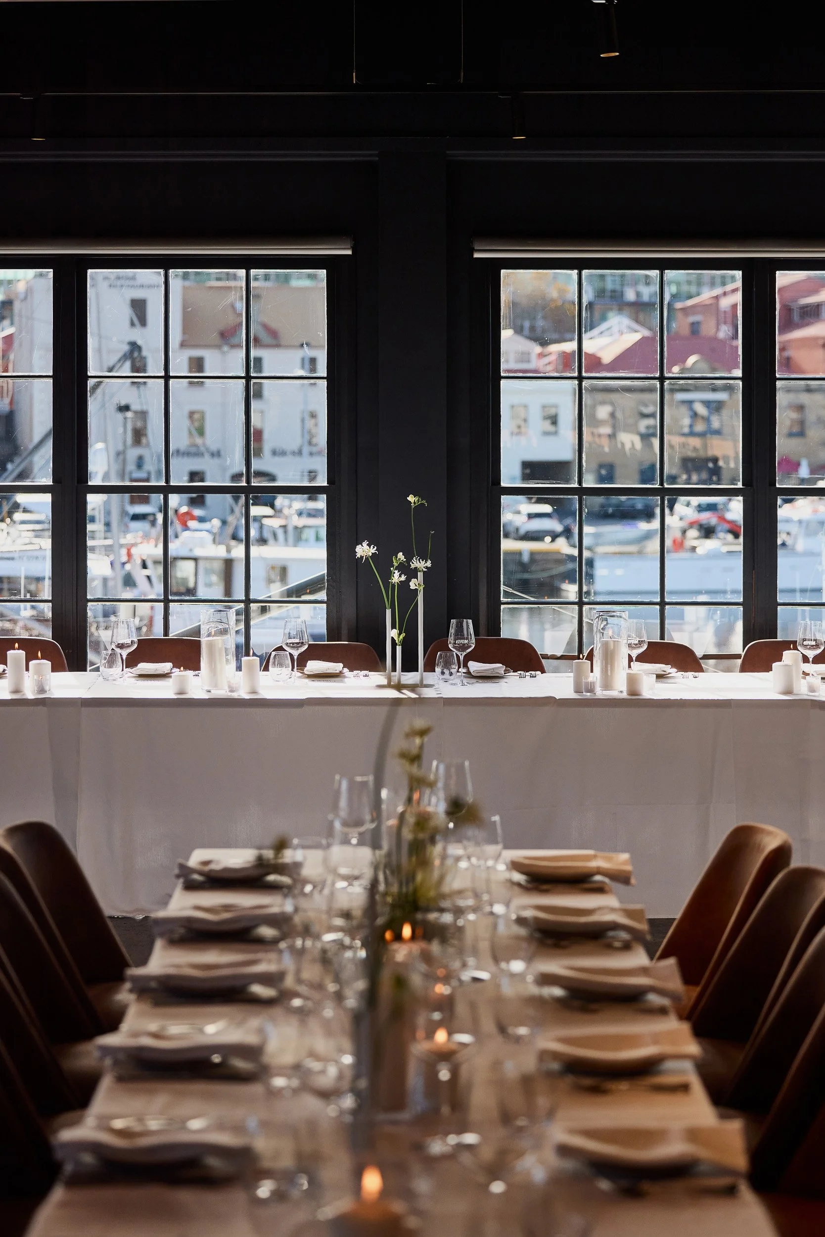 Interior of a restaurant with a long dining table set with plates, glasses, and candles, placed against large windows showing a view of city buildings outside.
