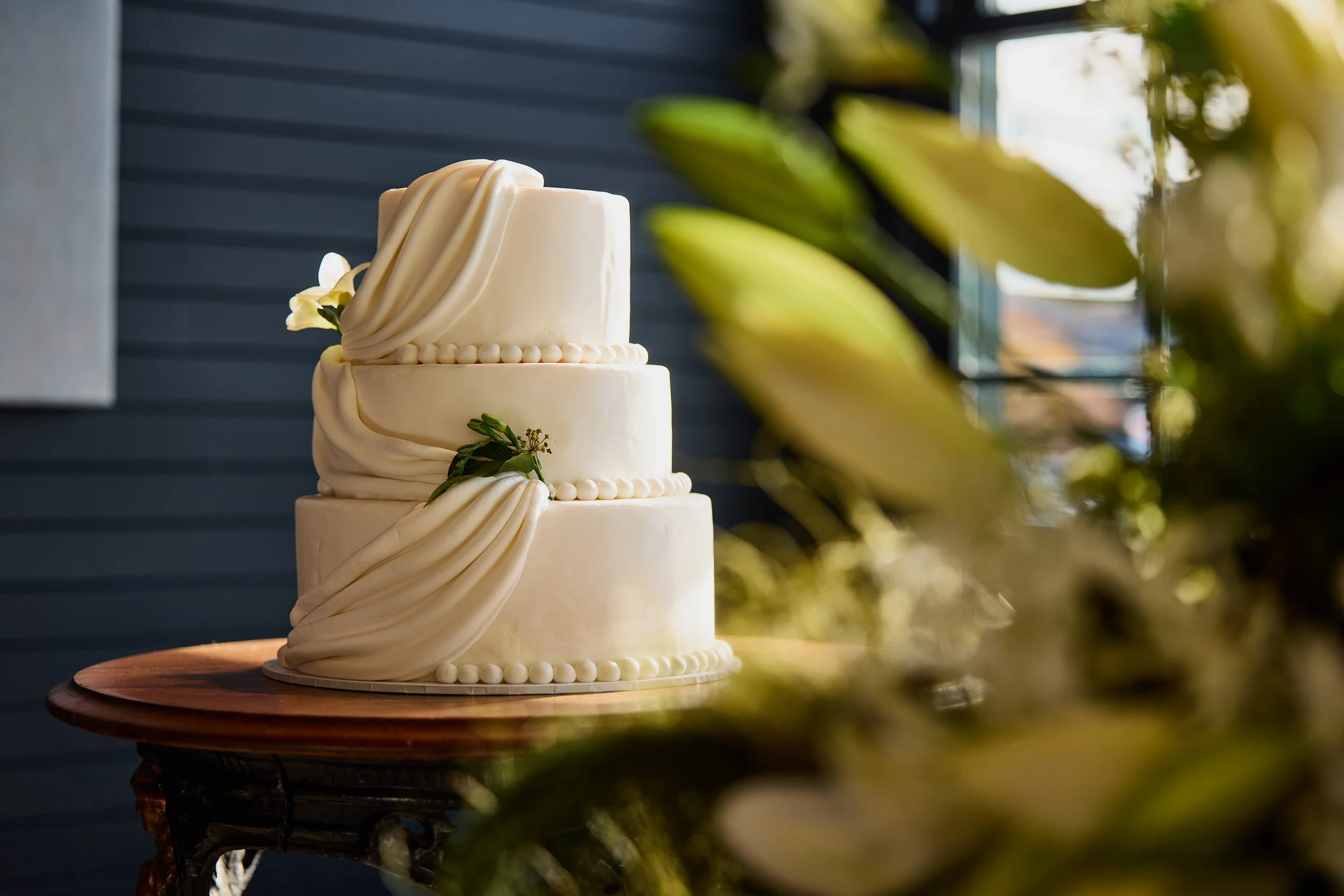 A three-tier white wedding cake decorated with draped fondant and small round pearls, adorned with white flowers and green foliage, placed on a round wooden cake stand.