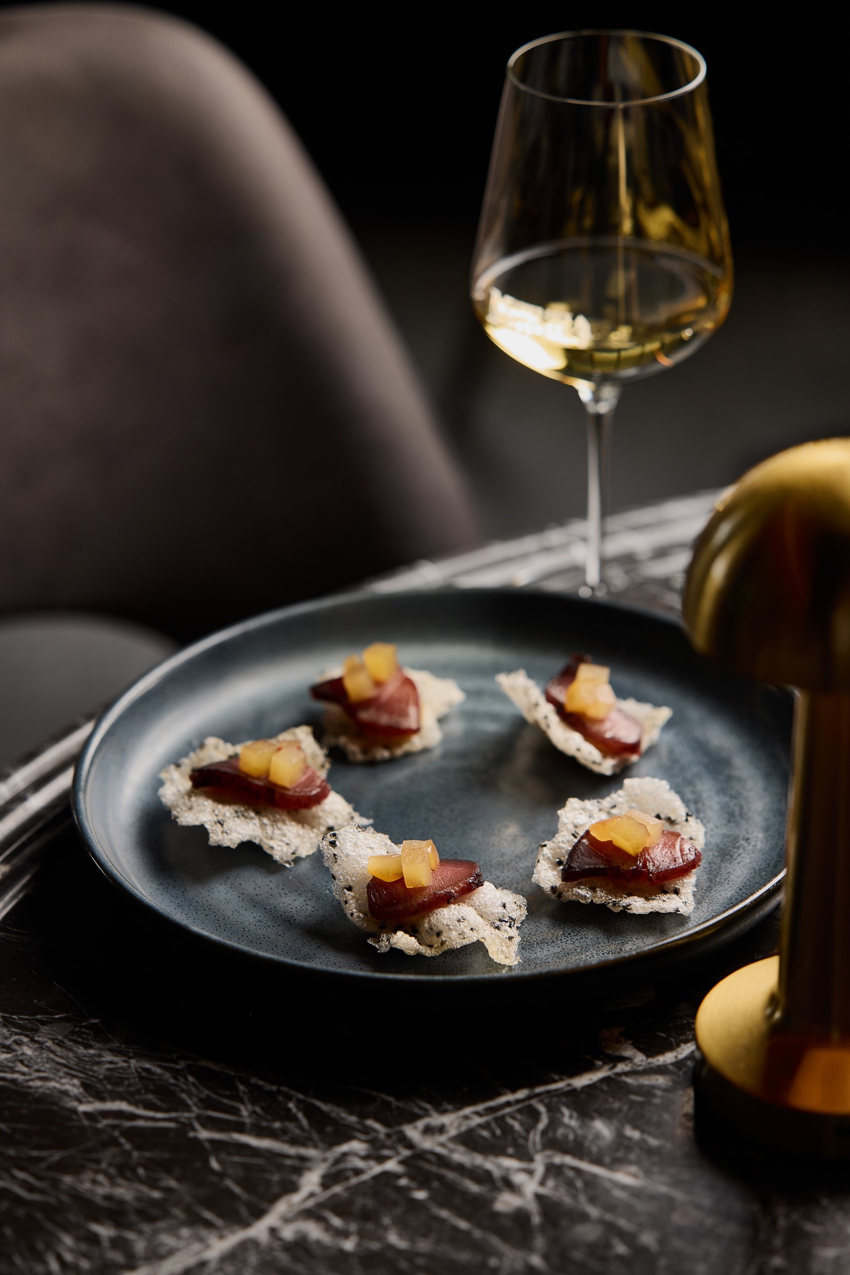 A glass of white wine and a plate with tuna appetizers on a dark marble table.