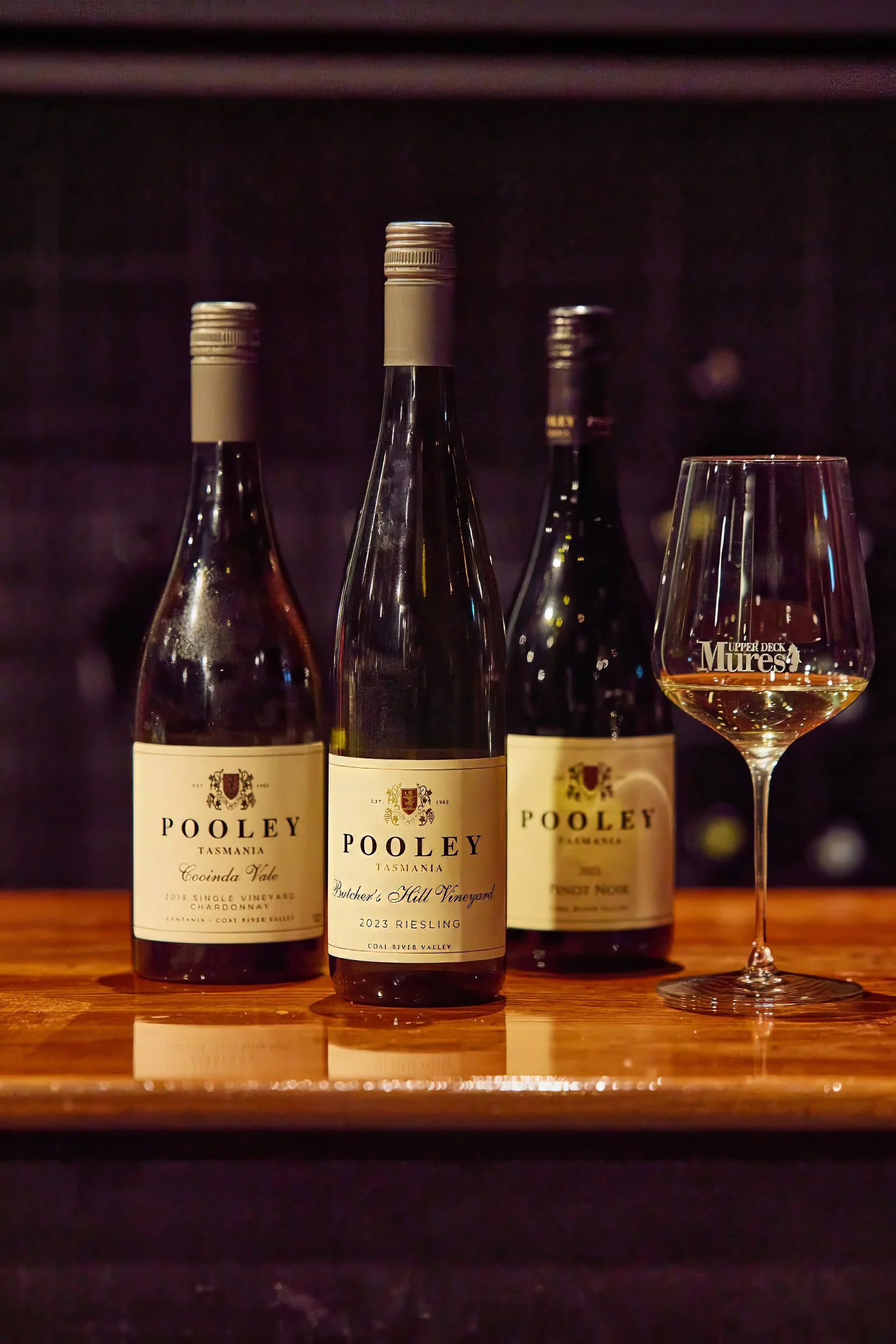 Three bottles of Pooley wine and a filled wine glass on a wooden surface, with a dark background.