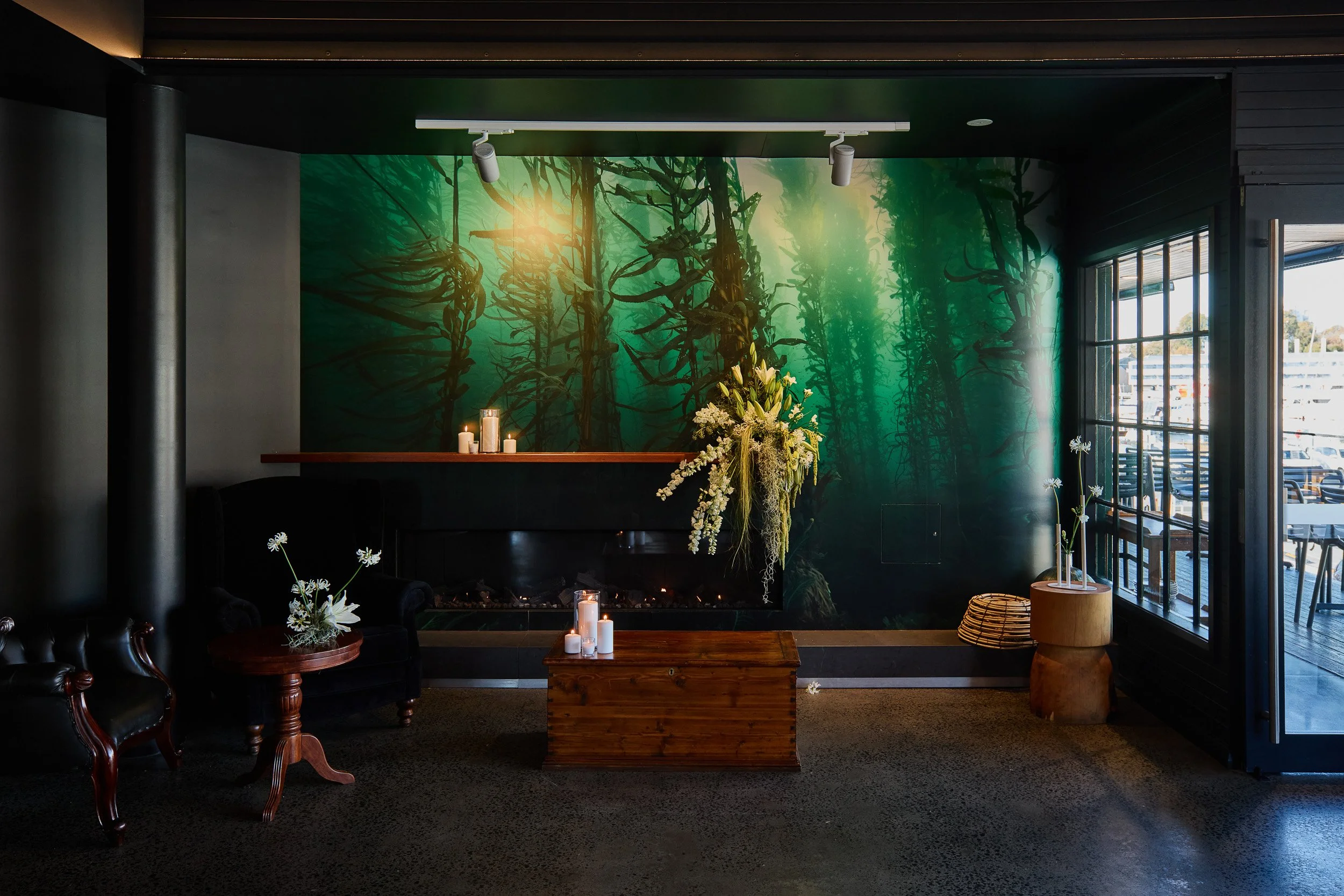 Interior of a modern room with a forest mural on the wall, candles on a fireplace mantle, a floral arrangement, a wooden table, upholstered chairs, and large windows showing a parking lot outside.