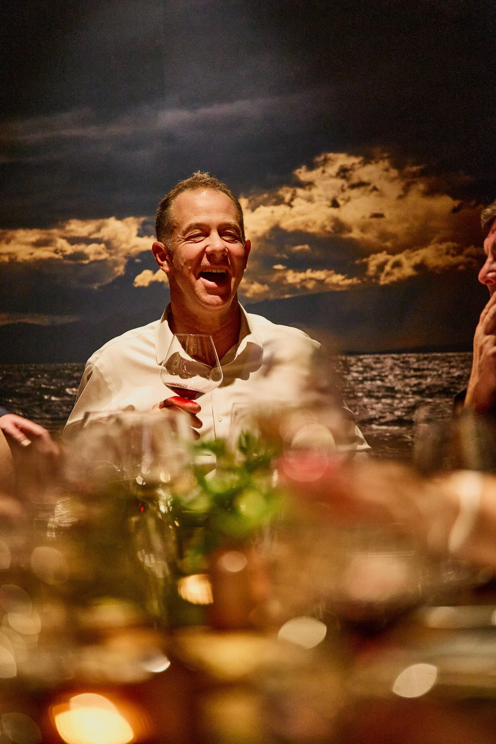 A man wearing a white shirt holding a glass of red wine, laughing and enjoying a social gathering at night with a cloudy sky and water in the background.
