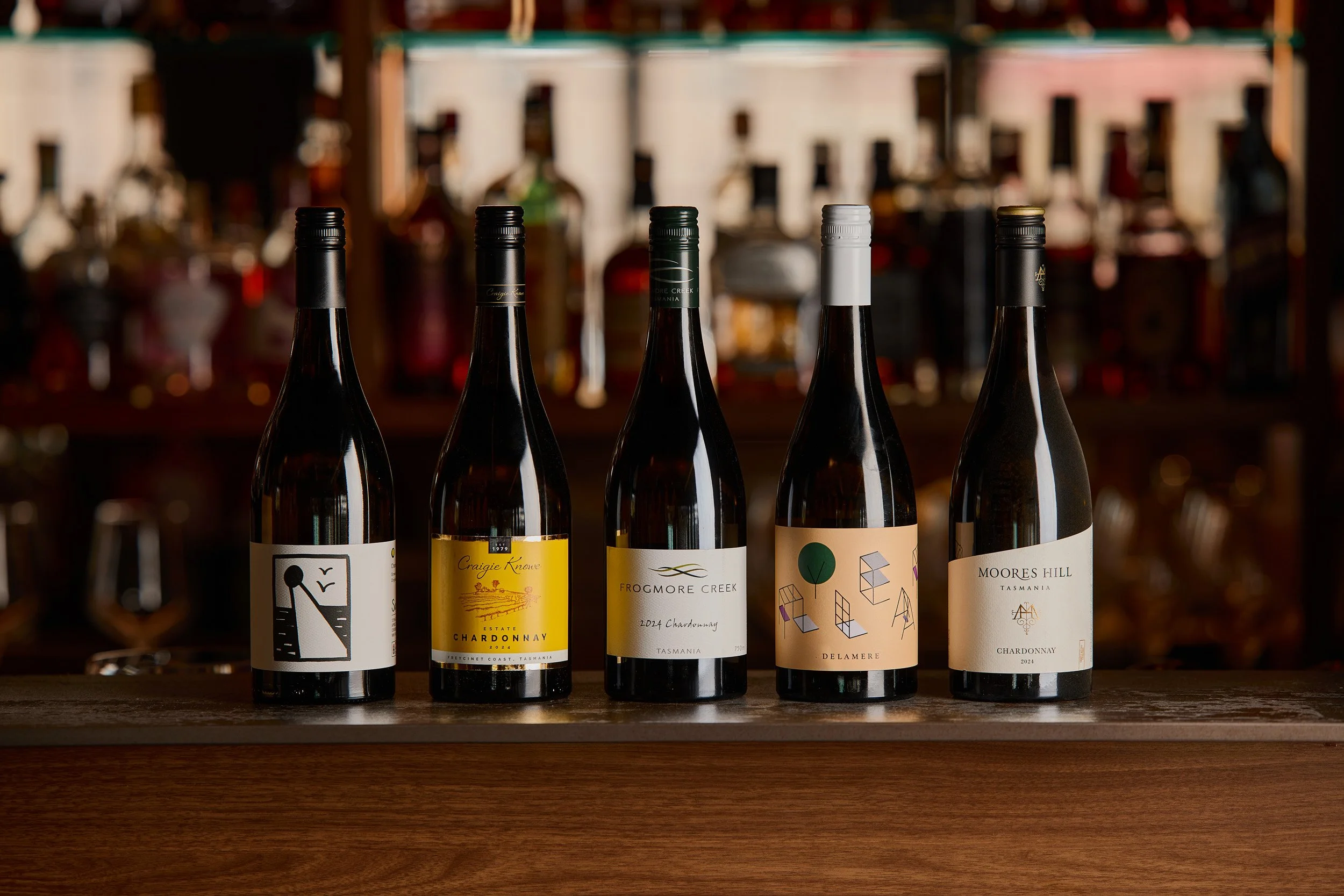 Several bottles of chardonnay wine on a wooden surface with blurred bottles and bar behind them.