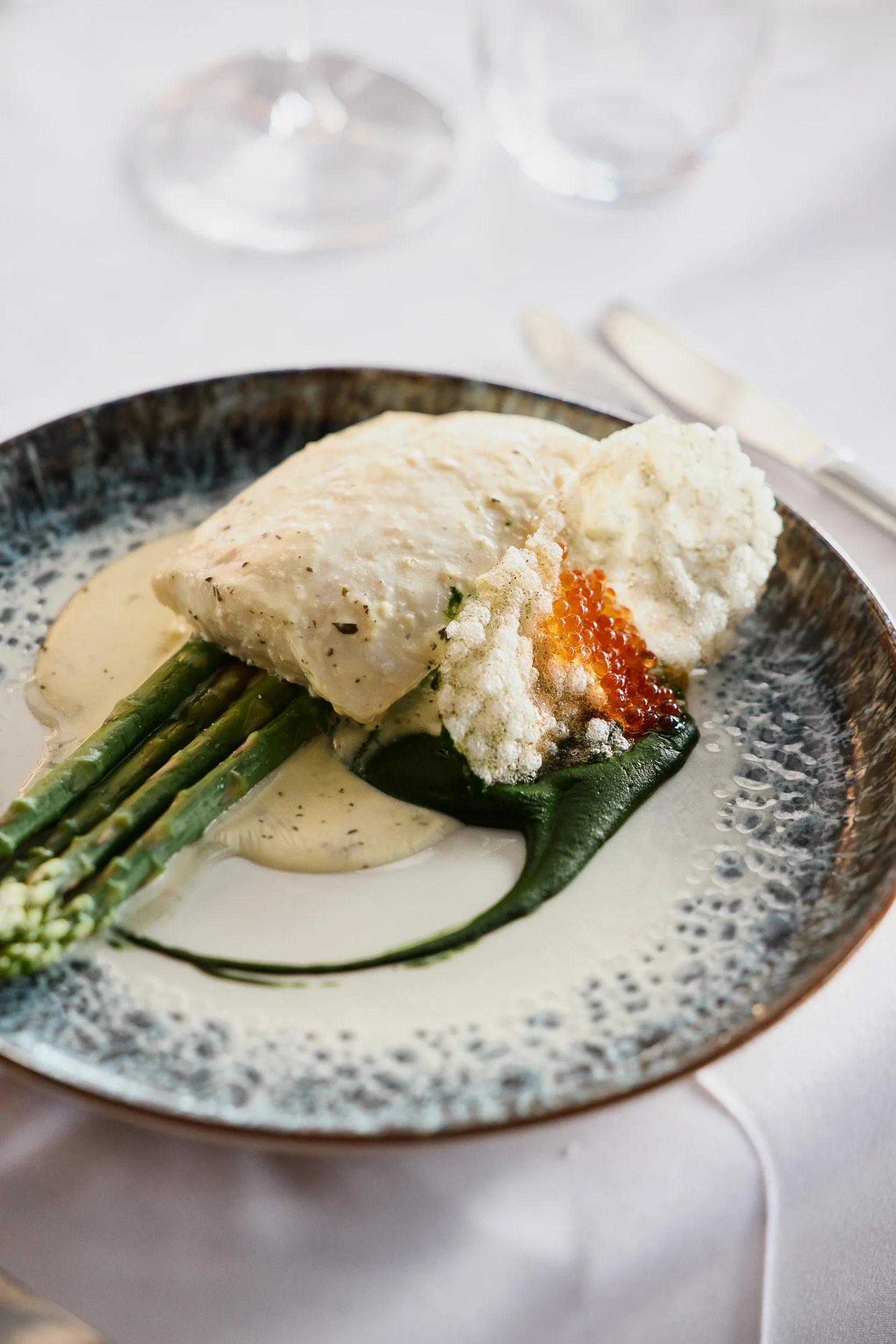 A plated meal featuring cooked white fish, asparagus spears, and a creamy sauce, garnished with small orange fish roe.