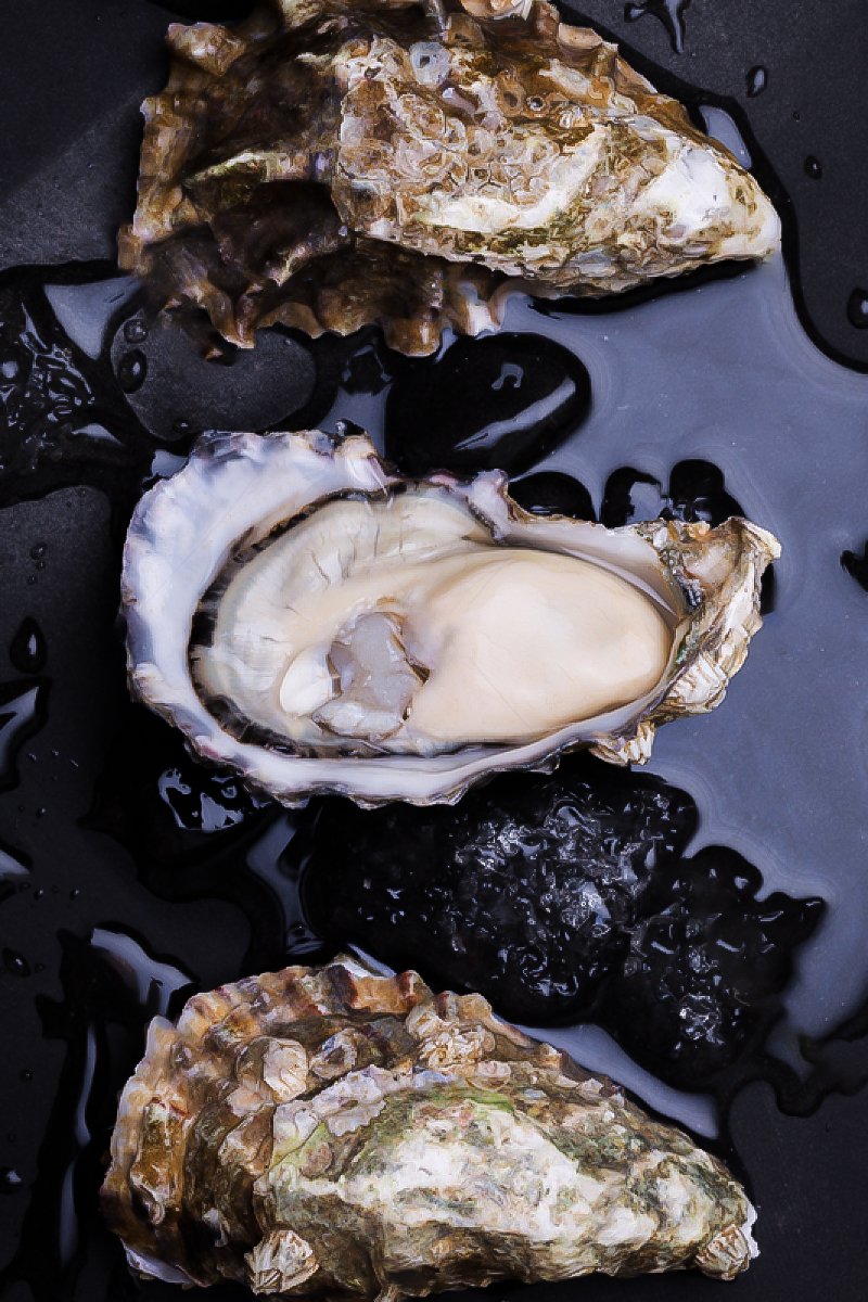 Three opened oysters on a black surface, some with shells still attached, and dark liquid surrounding them.