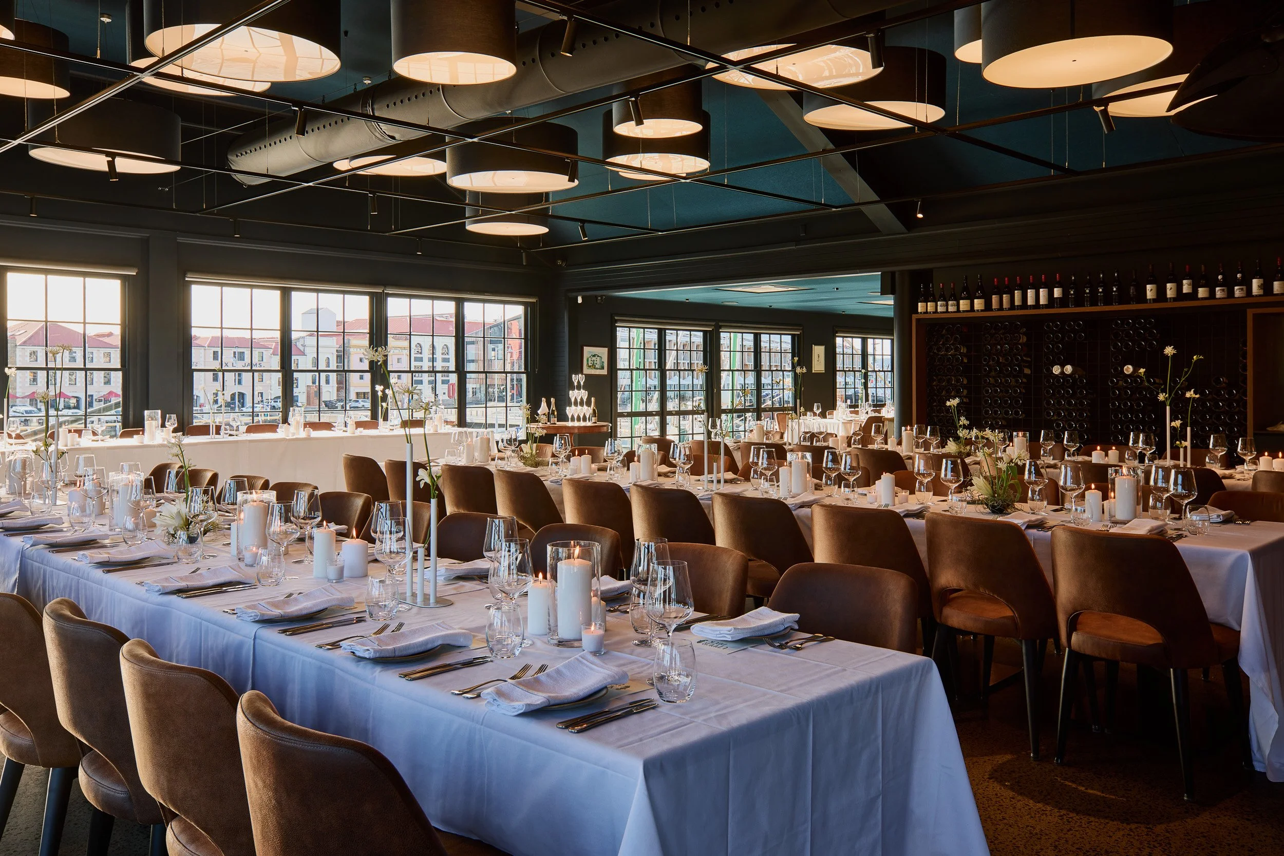 Elegant dining room set for a formal event with long tables covered in white tablecloths, decorated with candles and floral arrangements. Large windows let in natural light, showcasing a cityscape outside. The ceiling features modern circular light fixtures and dark hanging ductwork.