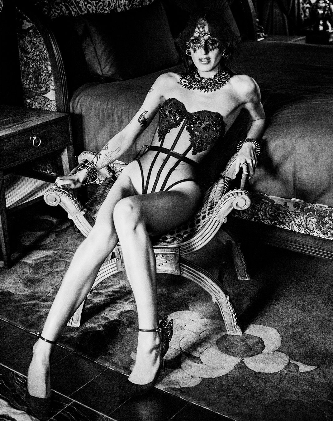 Cinematic black-and-white image of a model in intricate harness lingerie reclining on a deep red velvet sofa, captured by an acclaimed London editorial and fine-art photographer. Rich textures and dramatic mood – perfect for luxury fashion and beauty