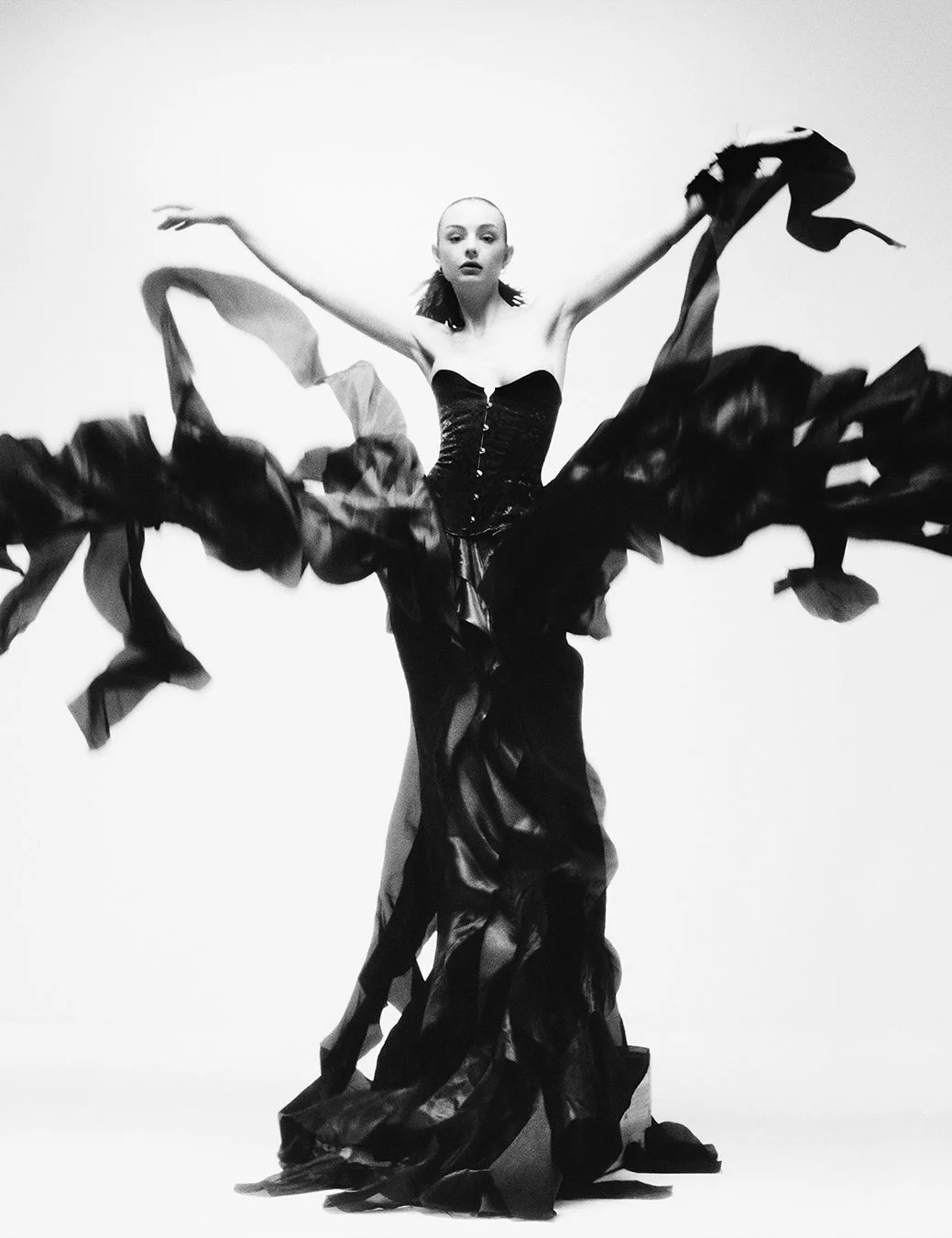 Dramatic black-and-white editorial shot of a model in a voluminous corseted gown with flowing fabric in motion, arms outstretched, photographed by a celebrated London fashion photographer. This worldwide-renowned commercial and editorial specialist e
