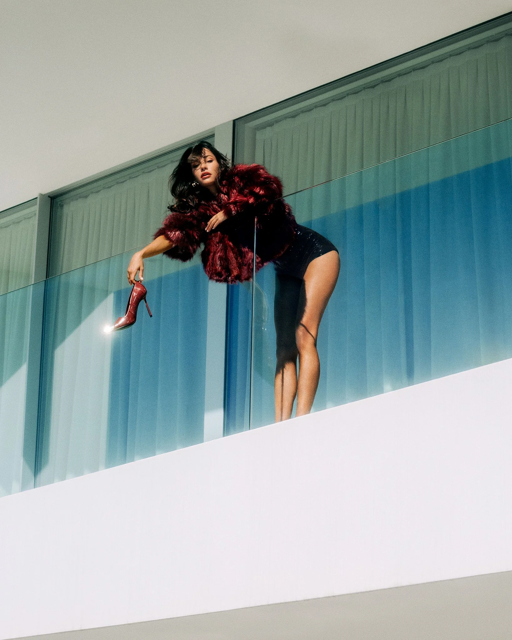 A model leans over a glass balcony railing in a red fur jacket, dangling a patent-leather heel from her fingertips — shot by London-based photographer Boris Edrosa