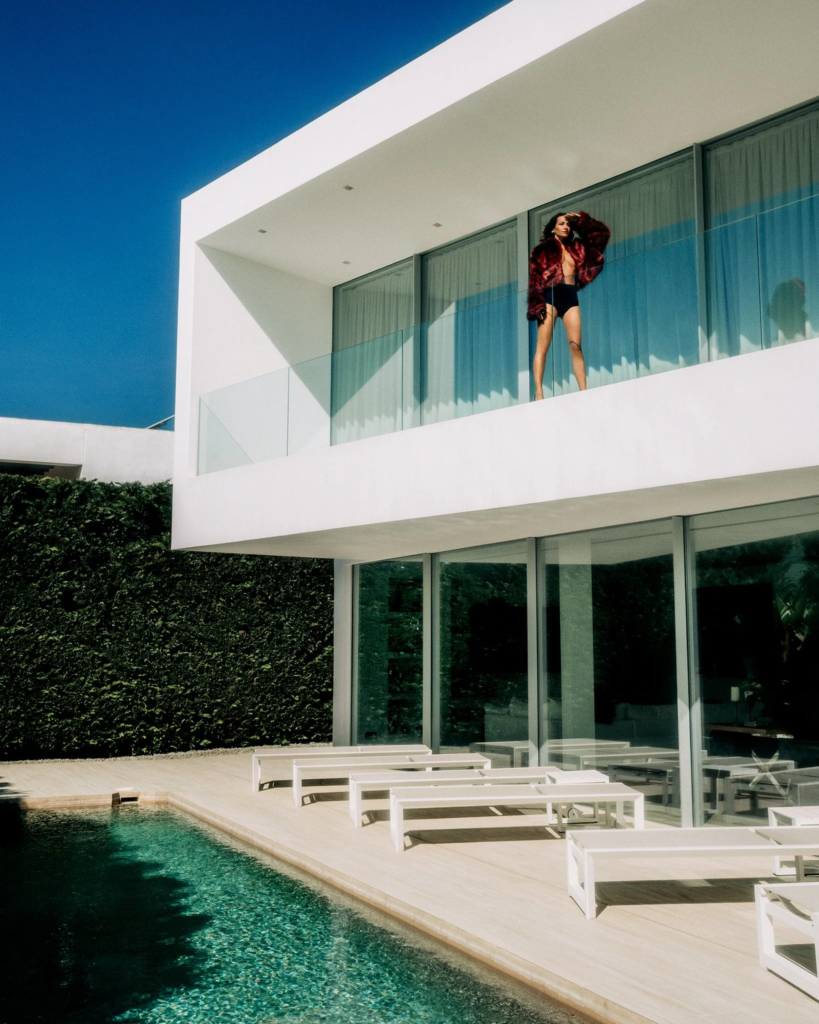 Model in a burgundy faux-fur jacket stands on a glass-balustraded balcony above an infinity pool, from an Ibiza Style Magazine editorial by fashion photographer Boris Edrosa