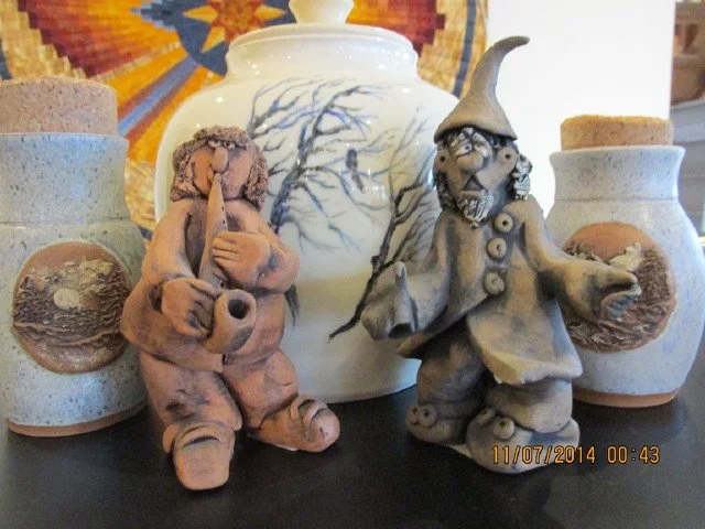  A figurine (left) playing a wind instrument beside a figurine of a wizard (right) displayed on a black surface in front of three vases, with a colourful wall ornament in the background 