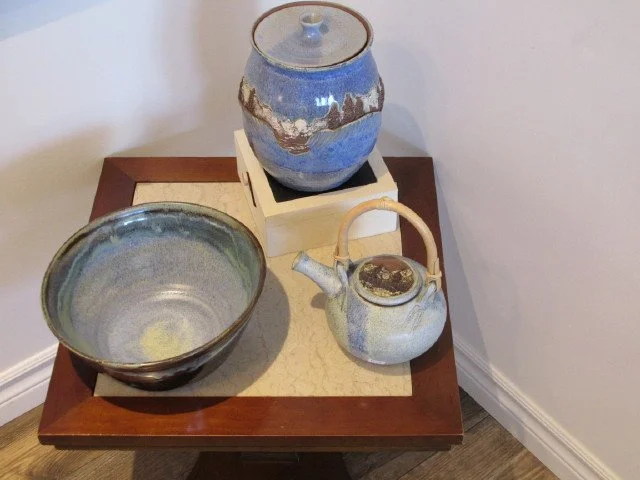  A bowl, stoneware container and tea kettle on a brown table with a light-brown mat in the center of the table 