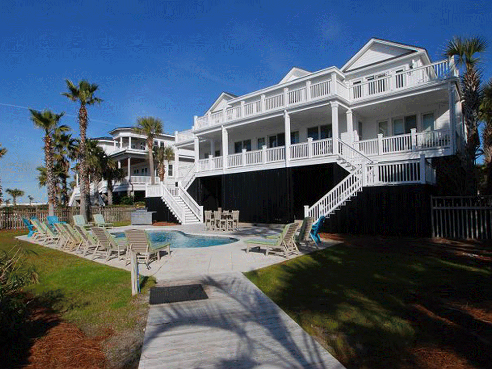 A large white beach house with multiple balconies, surrounded by palm trees, with a backyard pool area and lounge chairs, under a clear blue sky.