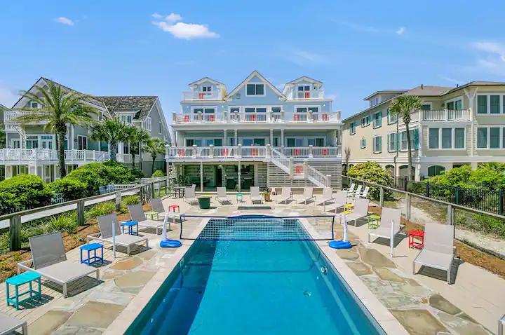Empty swimming pool with lounge chairs and small tables, in front of a large multi-story beach house with balconies, surrounded by palm trees and neighboring houses under a blue sky.