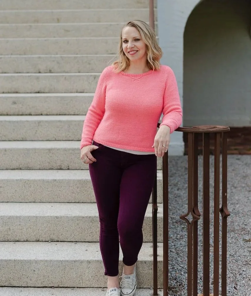 A woman with blonde hair wearing a pink sweater, purple pants, and white sneakers standing outdoors near stairs and a railing, smiling and looking away from the camera.