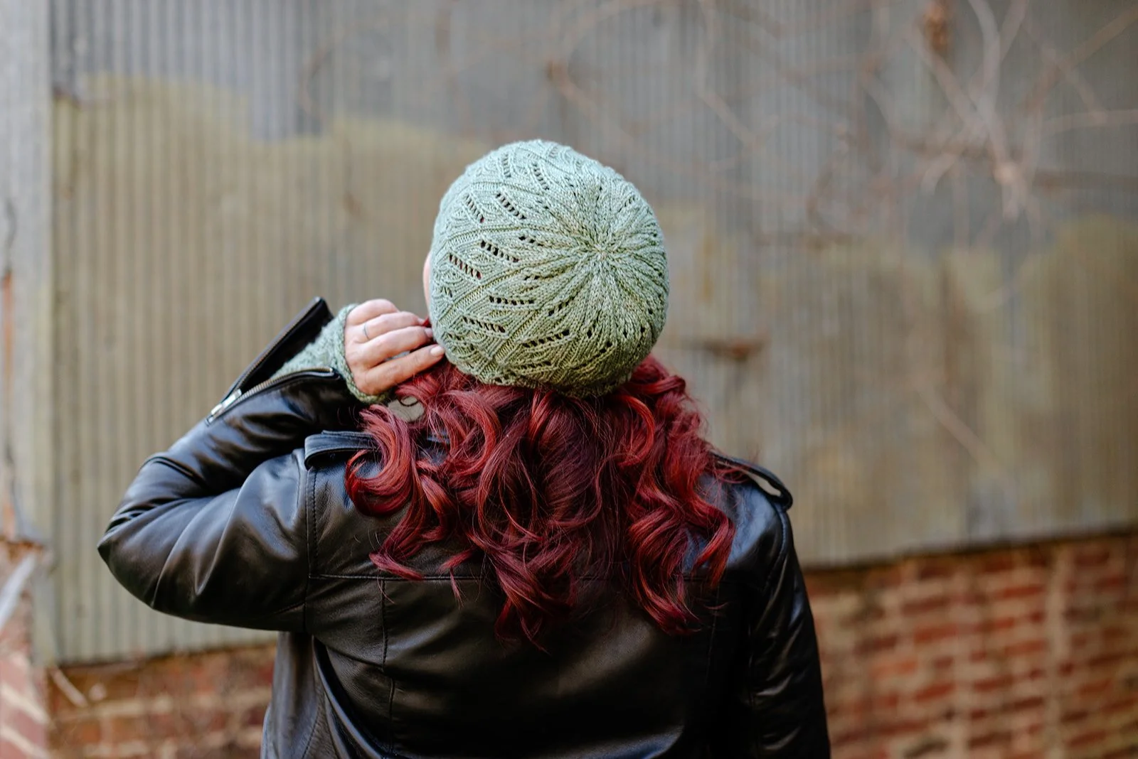 Back view of a woman with curly red hair wearing a gray knit hat and a black leather jacket, touching her ear, against a fence and brick wall background.