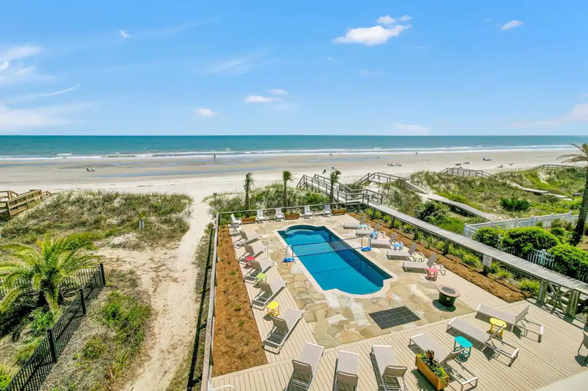 An outdoor pool area on a beachside property with lounge chairs, umbrellas, and a view of the sandy beach and ocean in the background.