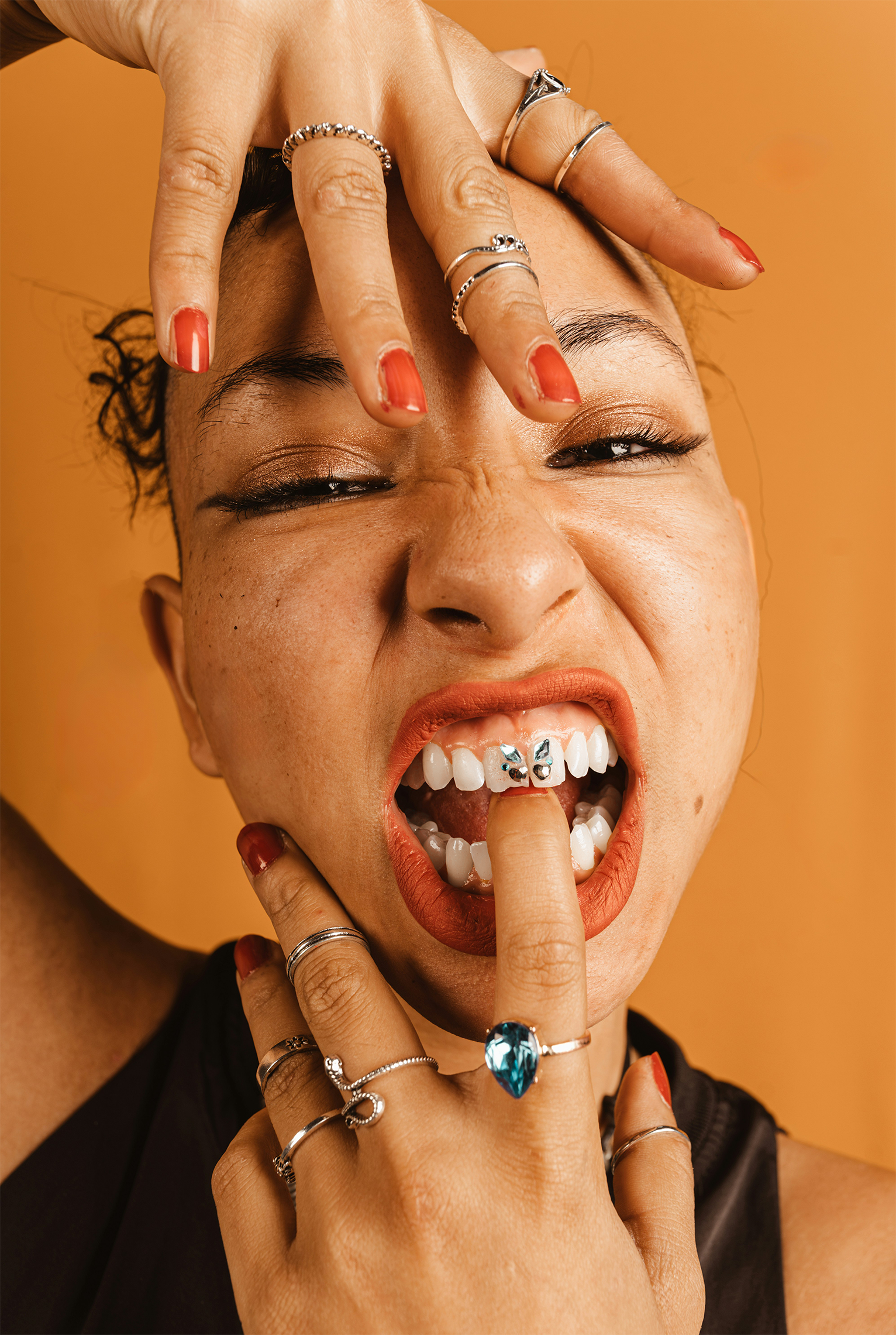 A woman with multiple rings and red nail polish is showing her teeth while licking her finger. She has a bright smile, worn makeup, and a vibrant expression.