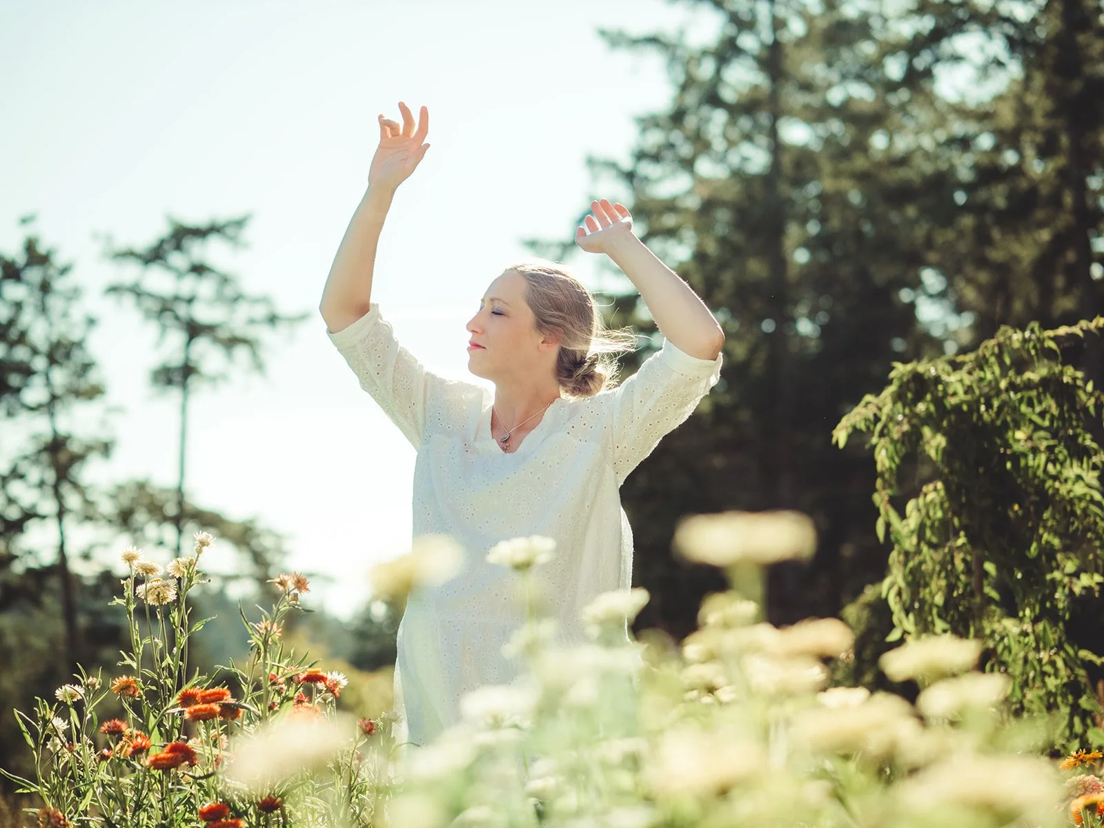 woman in white dress with eyes closed is standing in a field of wildflowers with her hands dancing in the air.