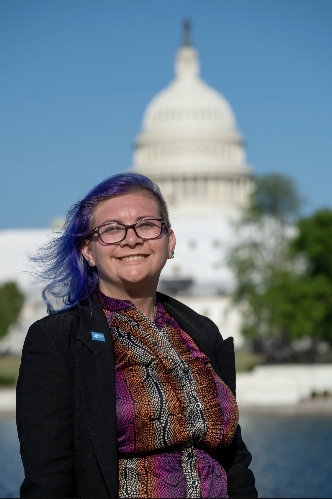A person with purple hair and glasses smiles in front of the U.S. Capitol building on a sunny day.