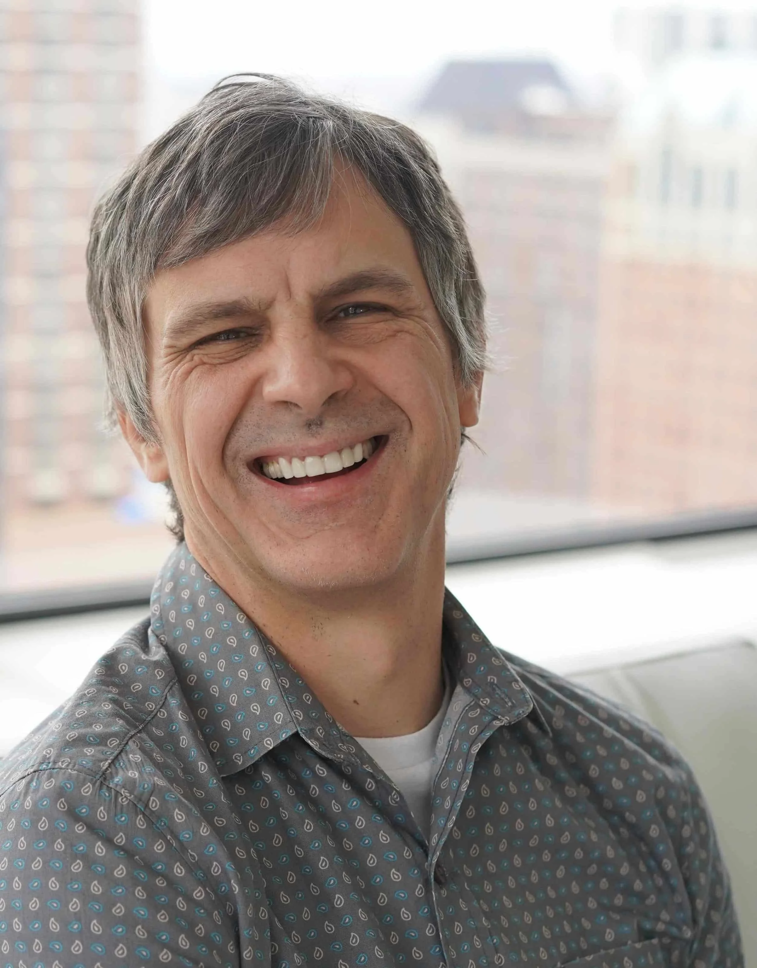 Man with gray hair smiling, wearing a patterned shirt, sitting indoors with a blurred cityscape in the background.
