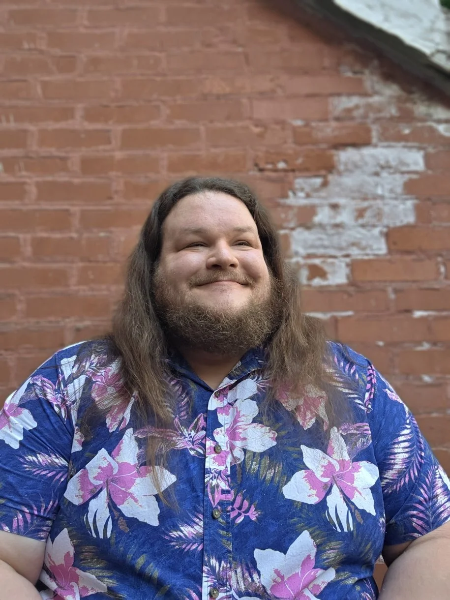 Smiling man with long hair and beard wears a blue floral shirt, standing in front of a weathered brick wall.