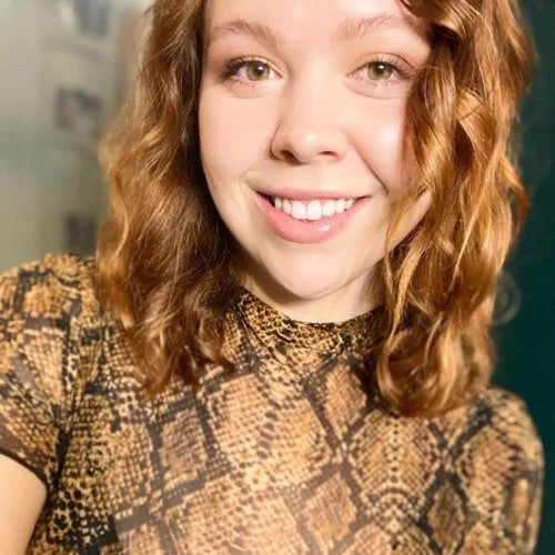 Smiling woman with wavy reddish-brown hair, wearing a brown snake-print top, posing for a headshot indoors.