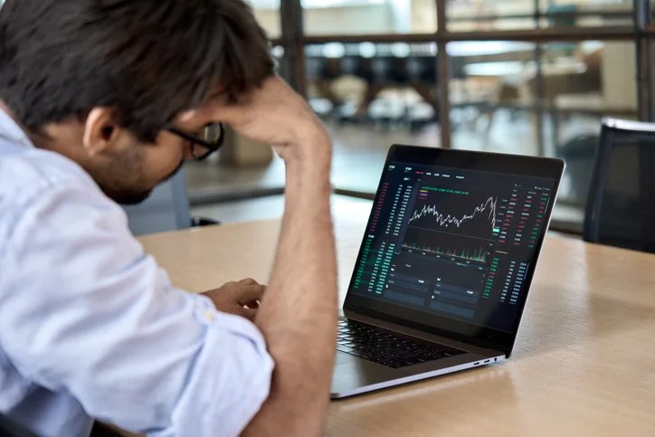 Man sitting at desk in corporate conference room, leaning his head in his hand, looking at financial charts on laptop