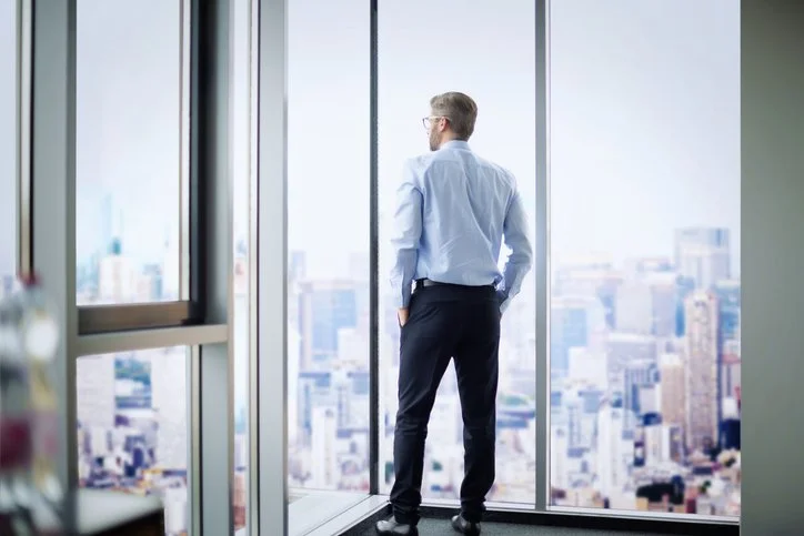 Man  with hands in pockets looking out window in an industrial office building, overlooking the city skyline