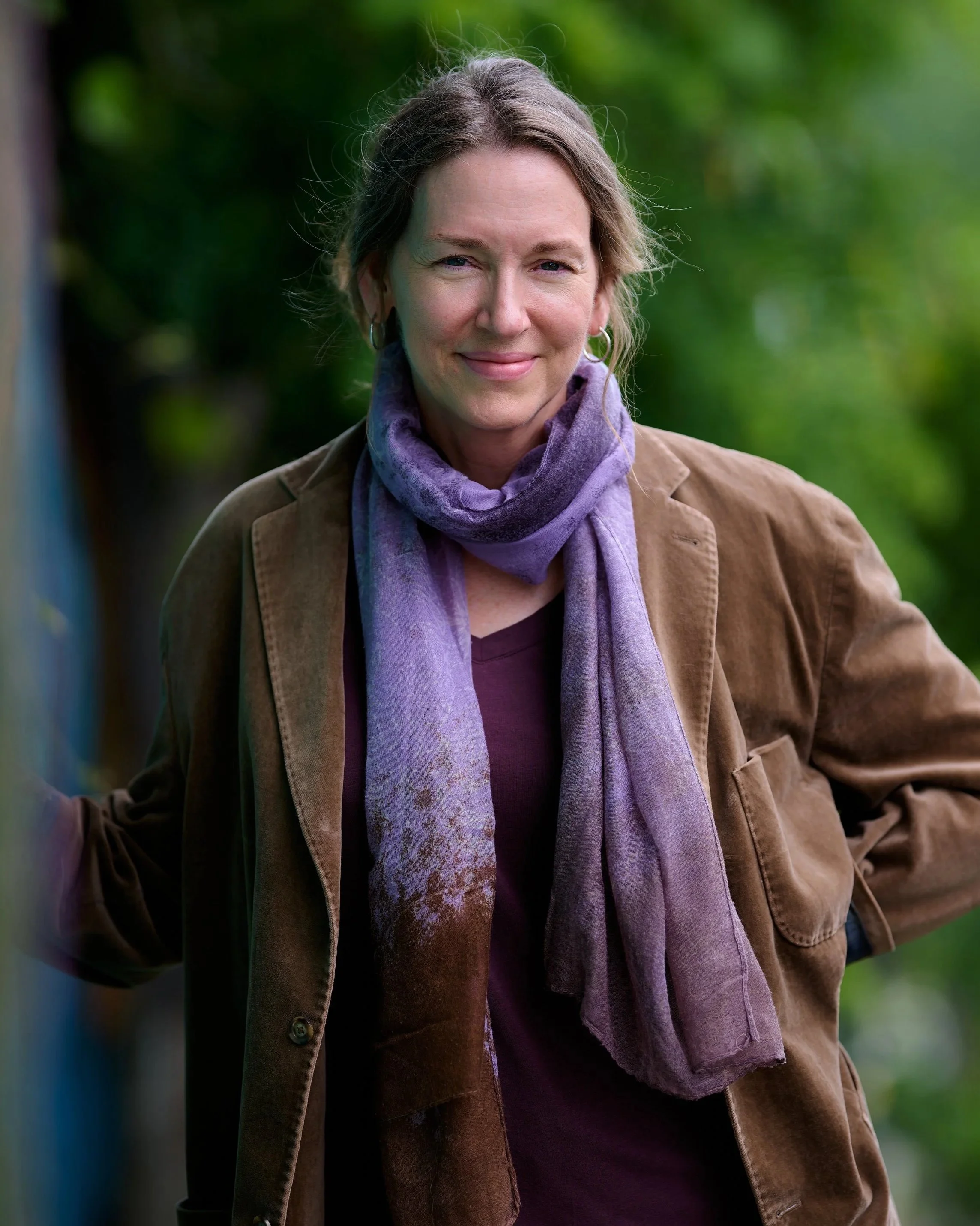 Close-up of a smiling woman with light brown hair, wearing a brown jacket and purple scarf, standing outdoors with a blurred green background.