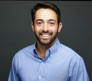 A man with dark hair and a beard wearing a light blue button-up shirt, smiling at the camera against a gray background.