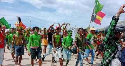 A photo of martial arts group members in Timor-Leste participating in a political campaign. They are walking down a street with flags and their bodies covered in green paint.