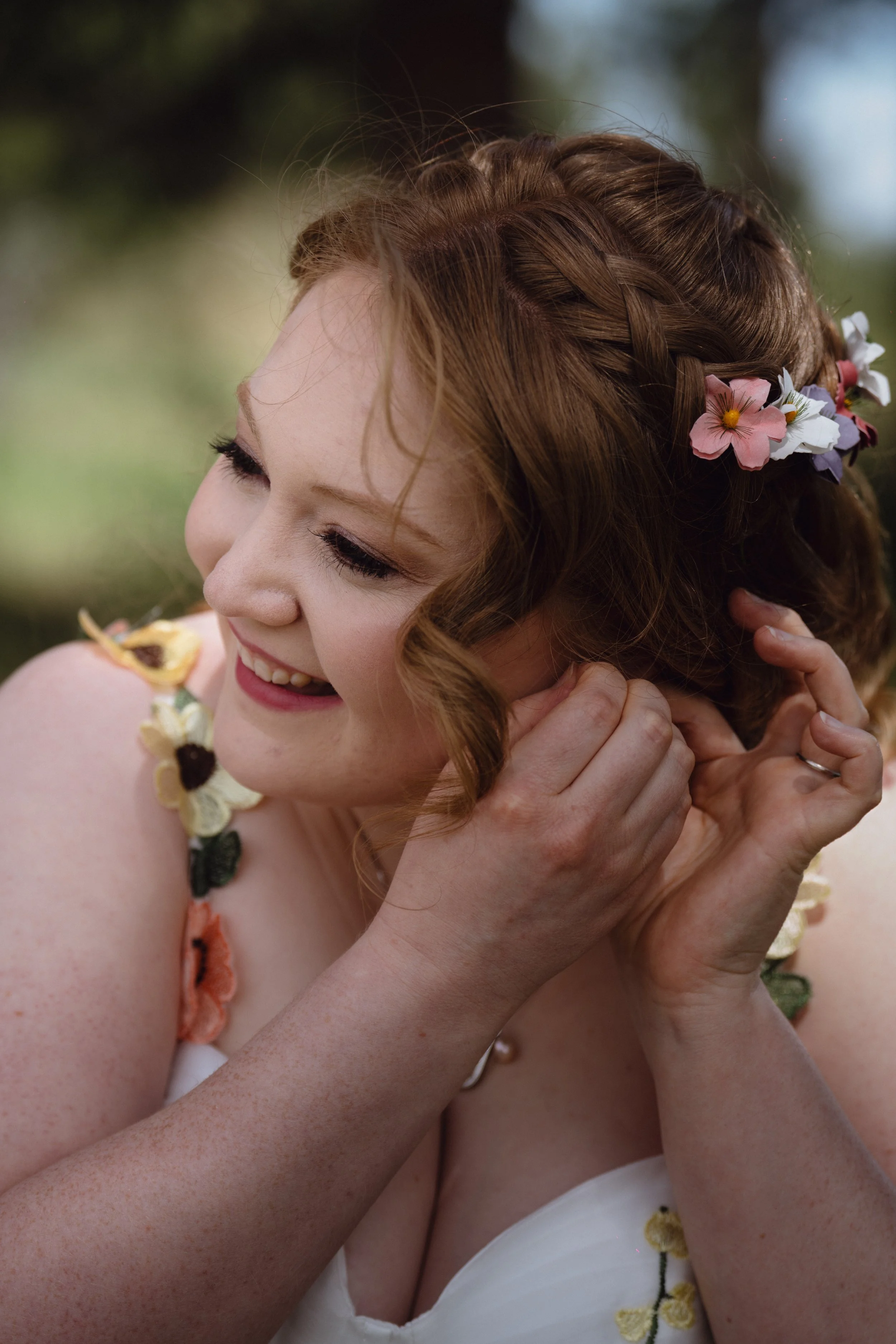 Close-up of a smiling bride with red hair, wearing floral accessories and a white dress, outdoors.