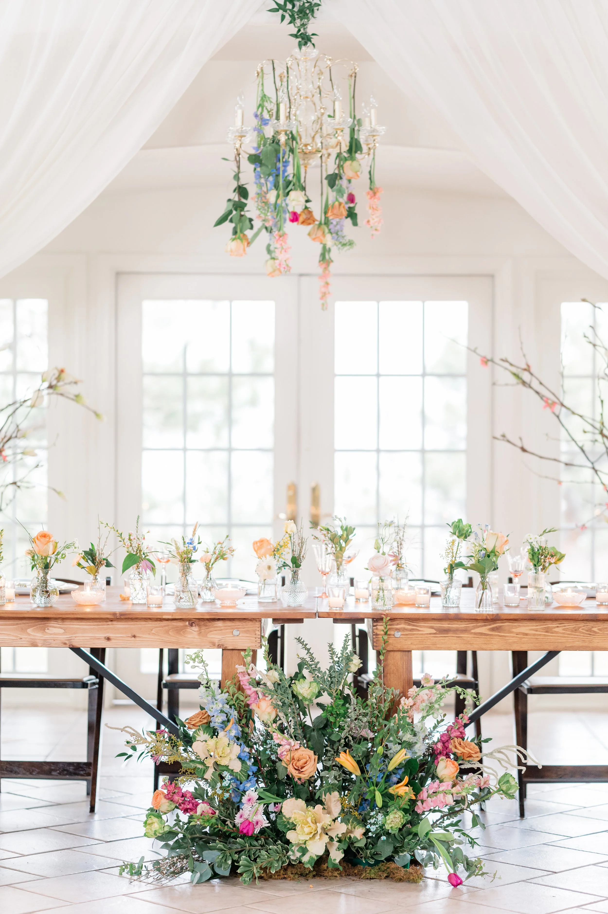 A wedding or event table decorated with a variety of colorful flowers, floral arrangements, and a chandelier with hanging flowers and candles in a bright room with large windows.