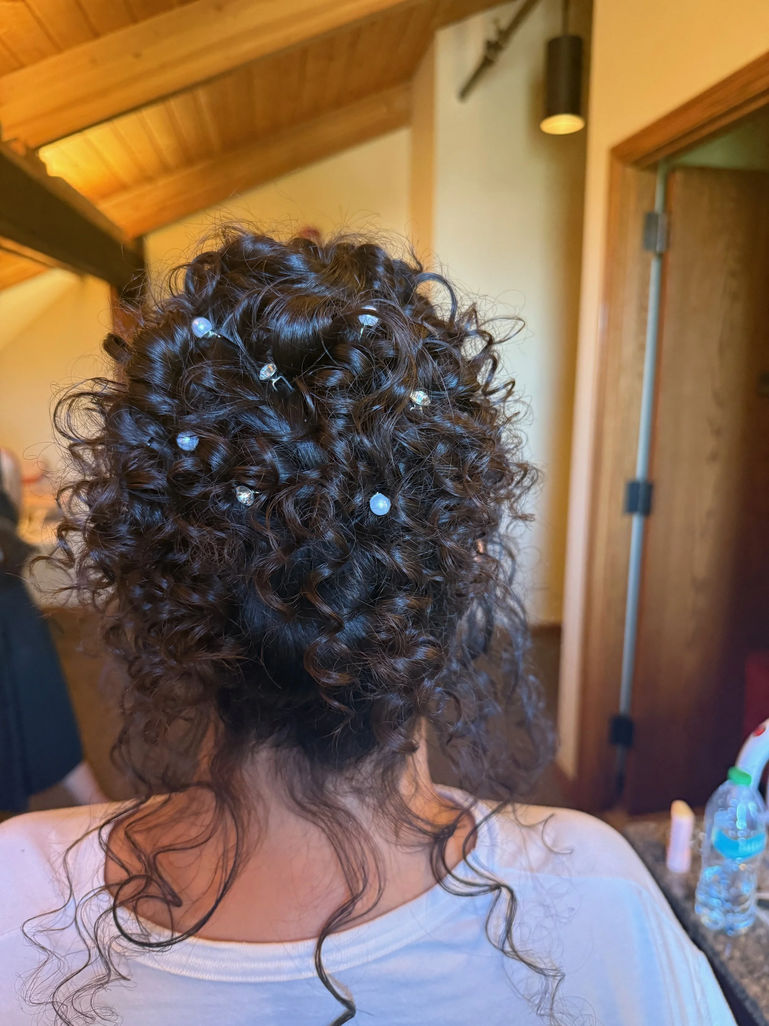 A bride with dark, curly hair decorated with small, shiny beads or clips, standing in a room with wooden ceiling beams and a doorway in the background.
