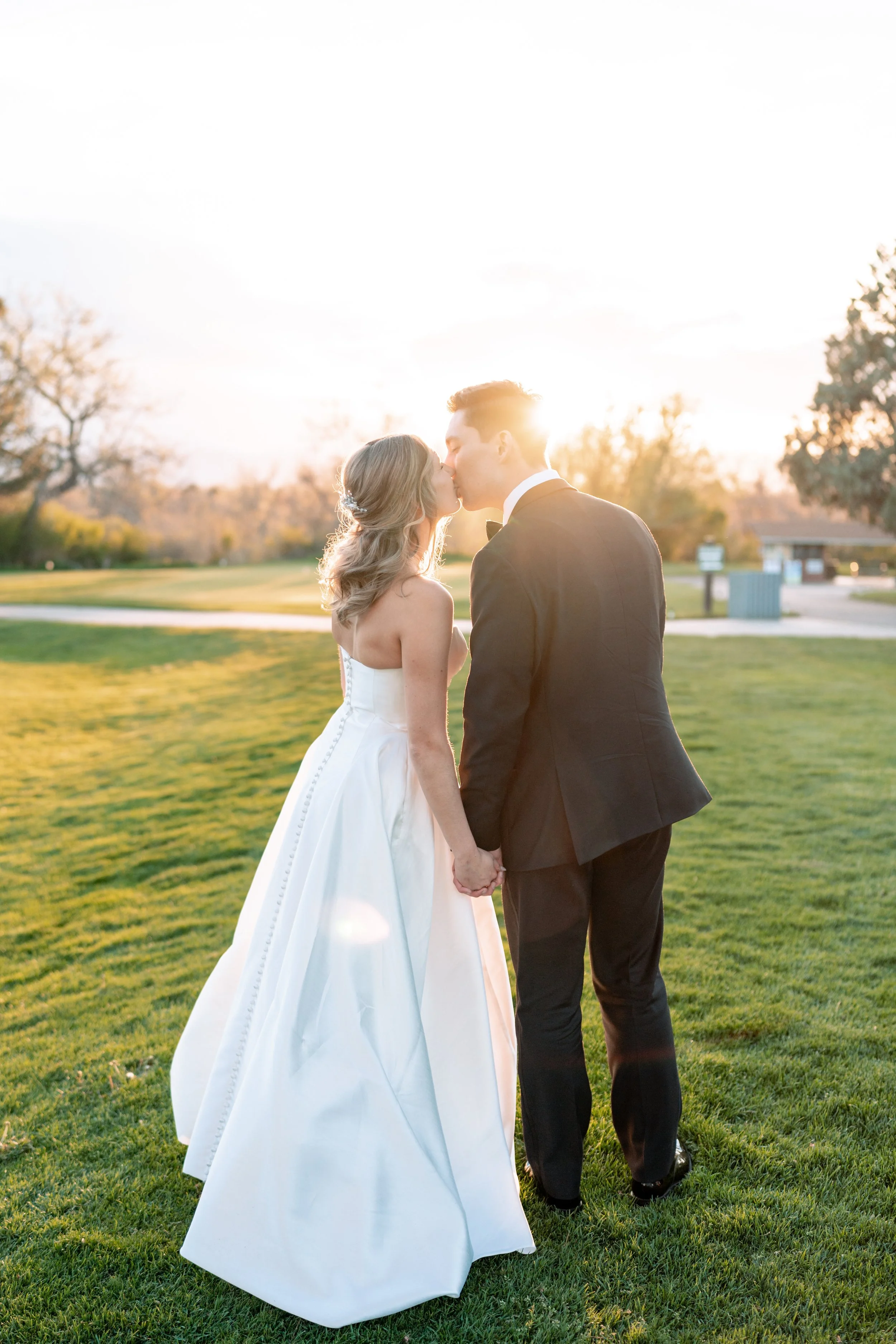 A bride and groom kissing outdoors at sunset, holding hands, with trees and a building in the background.