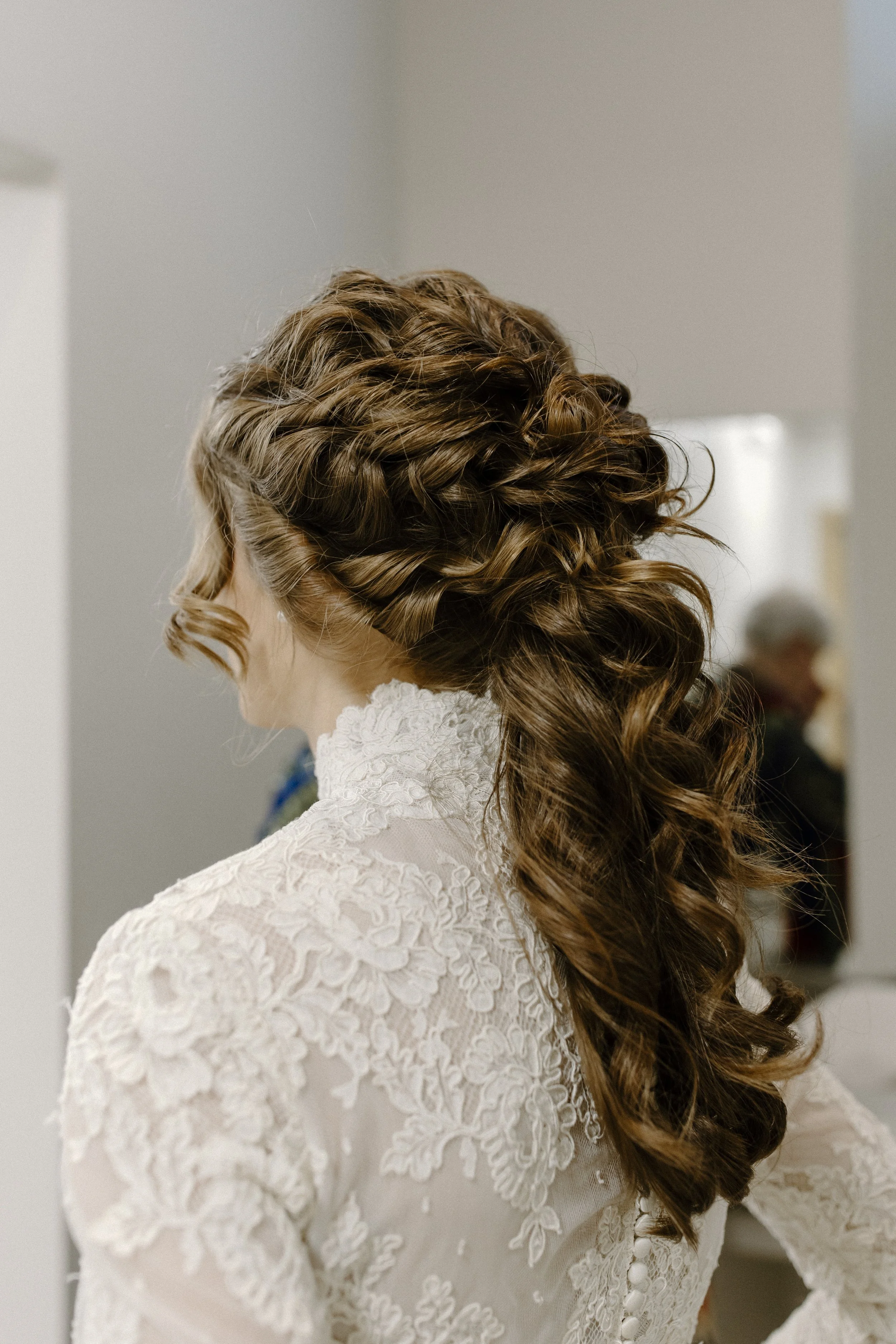 A bride with long, curly brown hair styled in an elegant updo with loose curls cascading down. She is wearing a white lace garment, possibly a wedding dress, with intricate floral embroidery and buttons in the back.