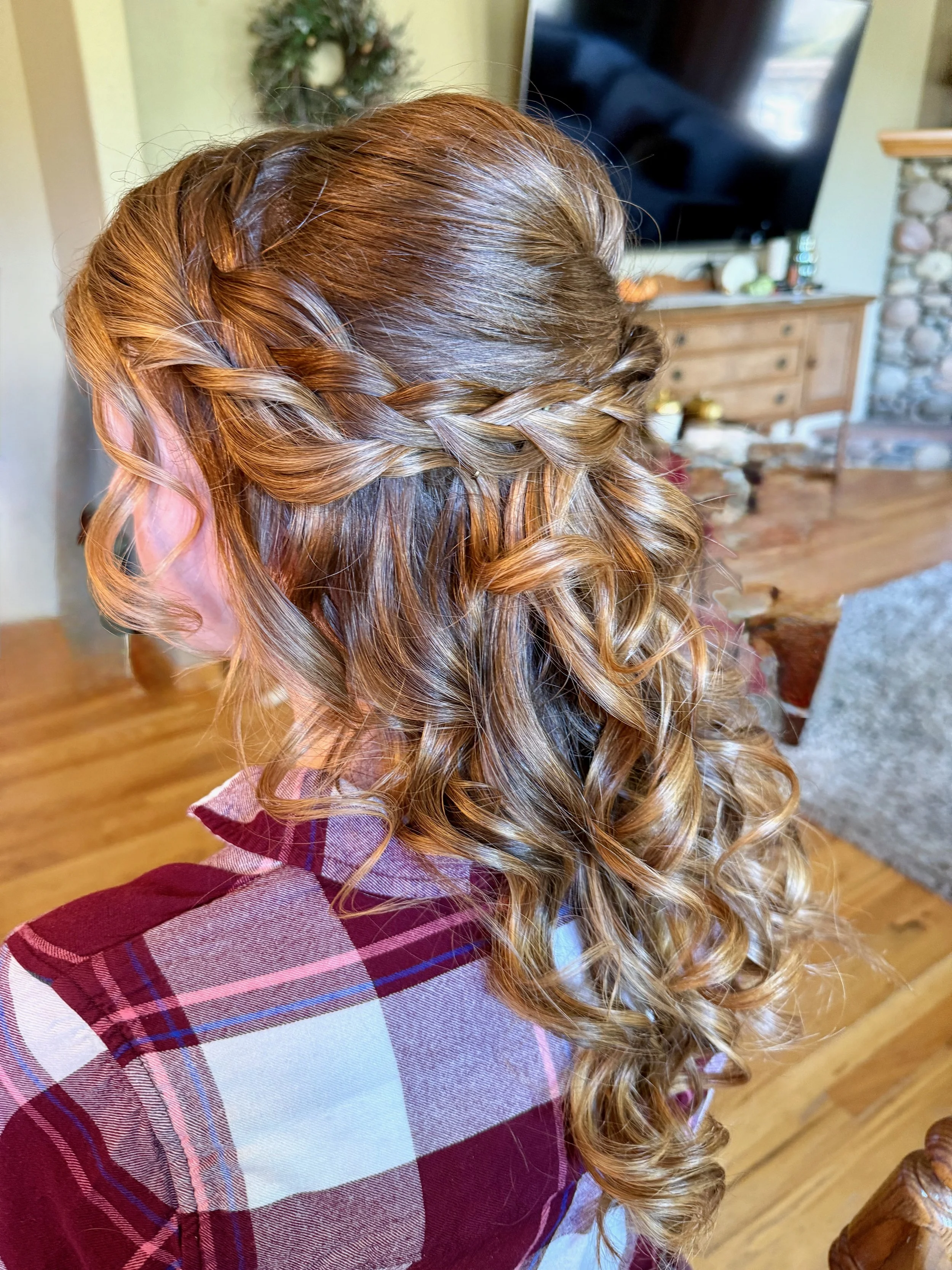 A bride with long, curly, reddish-blonde hair styled in a loose braid over her shoulder, wearing a plaid shirt with red, pink, purple, and white colors, sitting in a cozy living room with a TV, wooden furniture, and stone fireplace in the background.