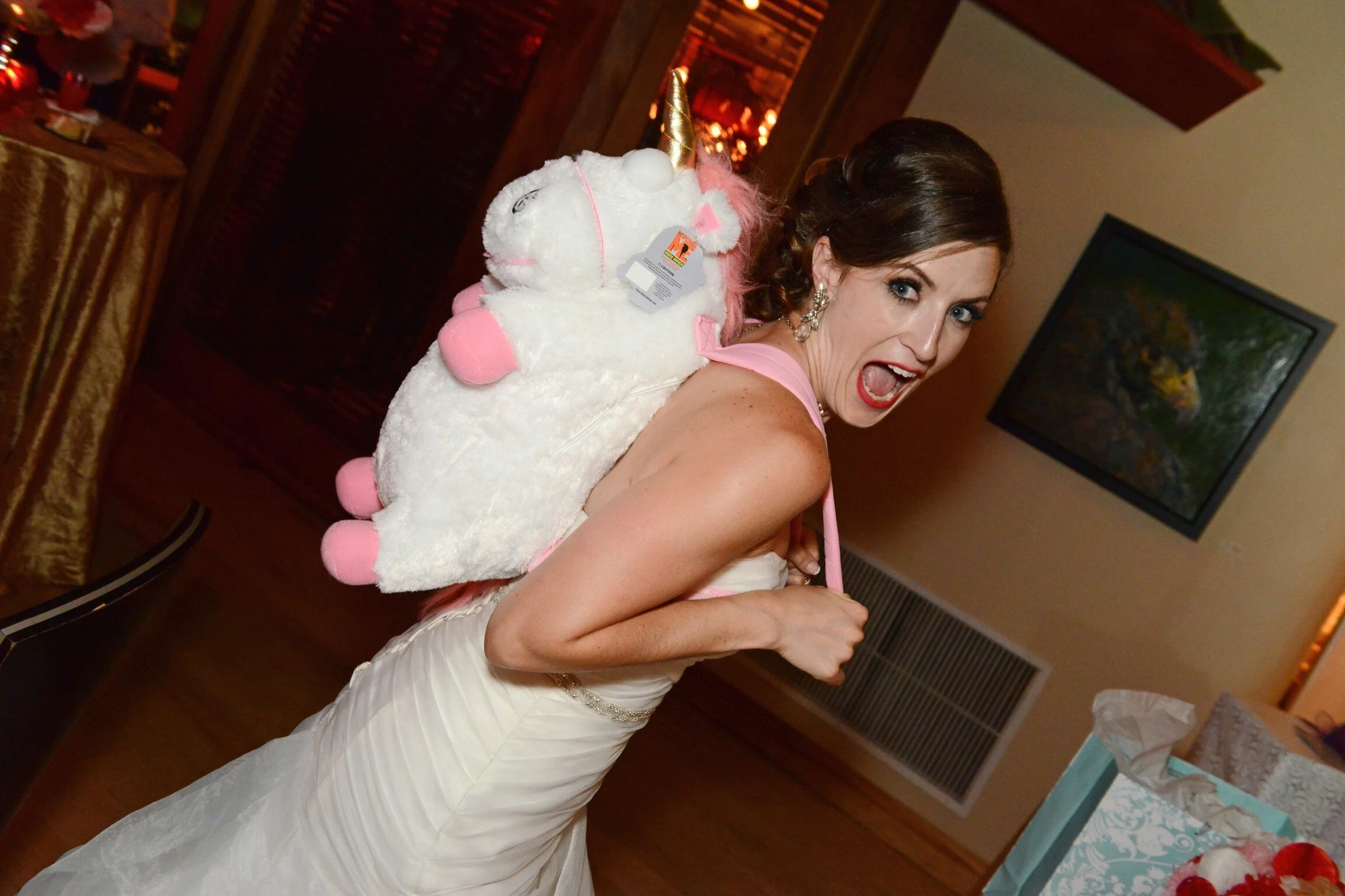 A woman in a white wedding dress with pink accents carries a white unicorn plush backpack with a golden horn and pink hooves, smiling with her mouth open at an indoor celebration or party.
