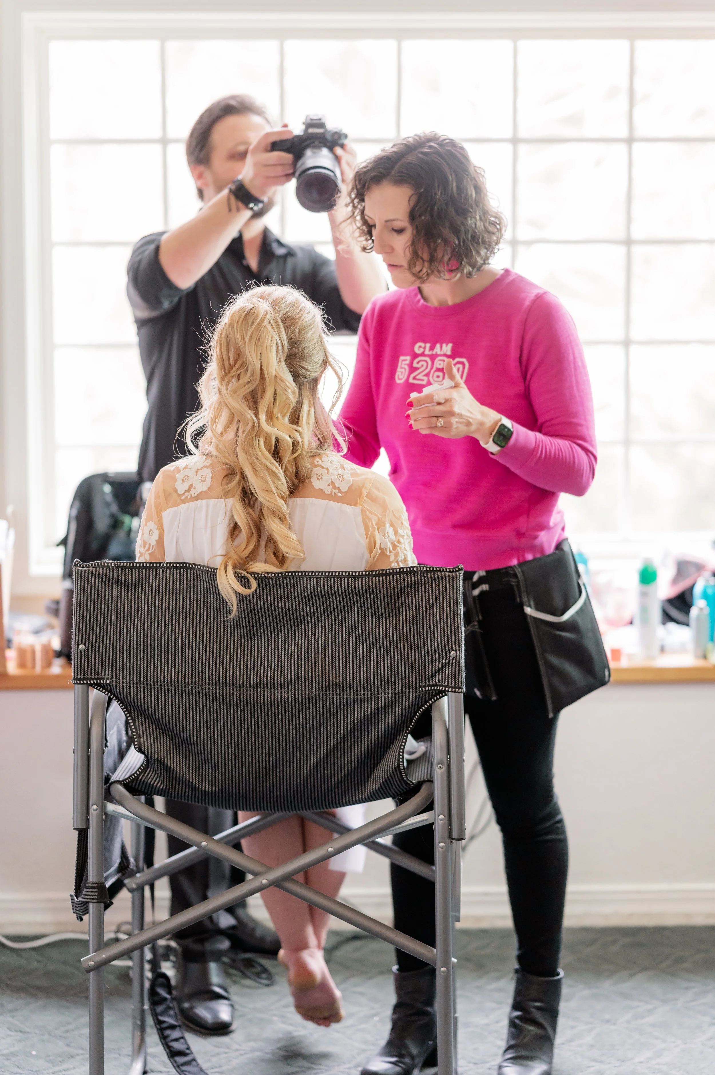 A woman with curly brown hair in a pink sweatshirt and a woman with long blonde hair in a white lace dress seated in a wheelchair. The woman in pink is pointing at the seated woman and looking down, while the woman in the wheelchair has her head slightly bowed. A photographer is standing behind them, taking a photo or video of the scene indoors with large windows in the background.